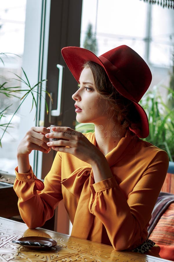 Ragazza in un caffè con una tazza di caffè e un cappello Ritratto della ragazza sensuale che porta cappello e blusa flosci con l' fotografia stock Ragazza in un caffè con una tazza di caffè e un cappello Ritratto della ragazza sensuale che porta cappello e blusa flosci con l' fotografia stock