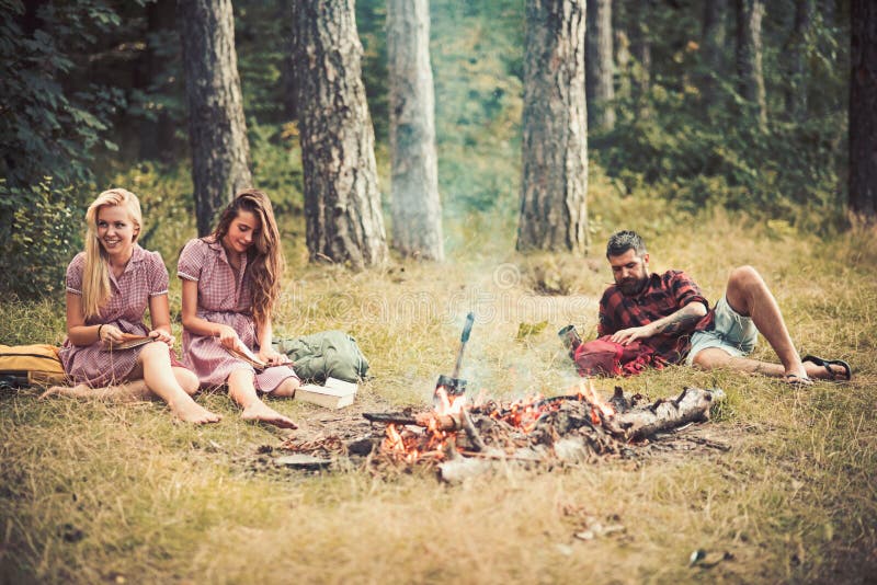 Ragazze che si siedono accanto al fuoco di accampamento mentre libri di lettura Tipo barbuto che si trova sull'erba negli amici d fotografia stock libera da diritti Ragazze che si siedono accanto al fuoco di accampamento mentre libri di lettura Tipo barbuto che si trova sull'erba negli amici d fotografia stock libera da diritti