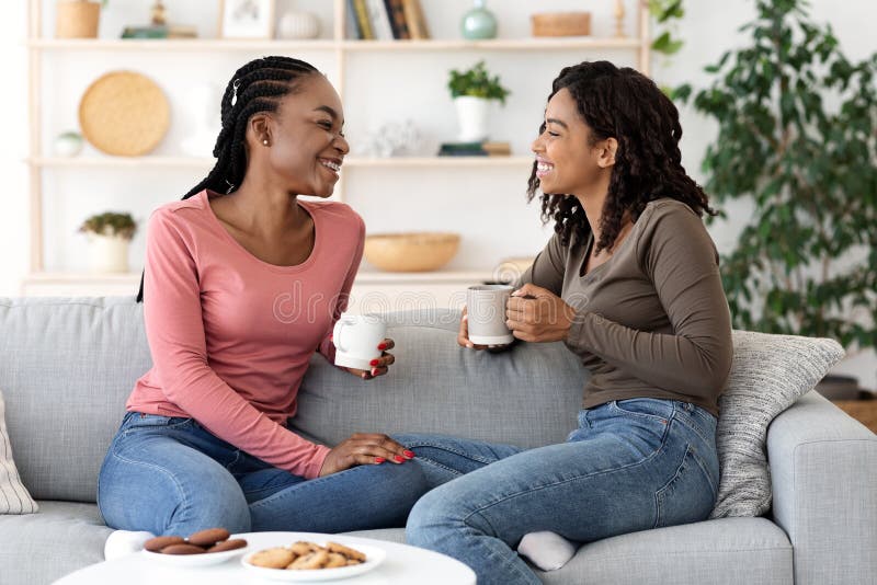 Black girlfriends enjoying weekend together, drinking coffee and talking on couch at home. Black girlfriends enjoying weekend together, drinking coffee and talking on couch at home