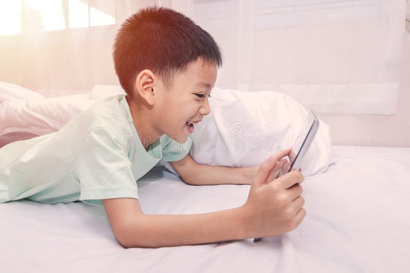 Ragazzino che guarda una tavoletta e gioca mentre sta sdraiato sul letto a casa. bambino che usa dispositivi intelligenti online c immagine stock