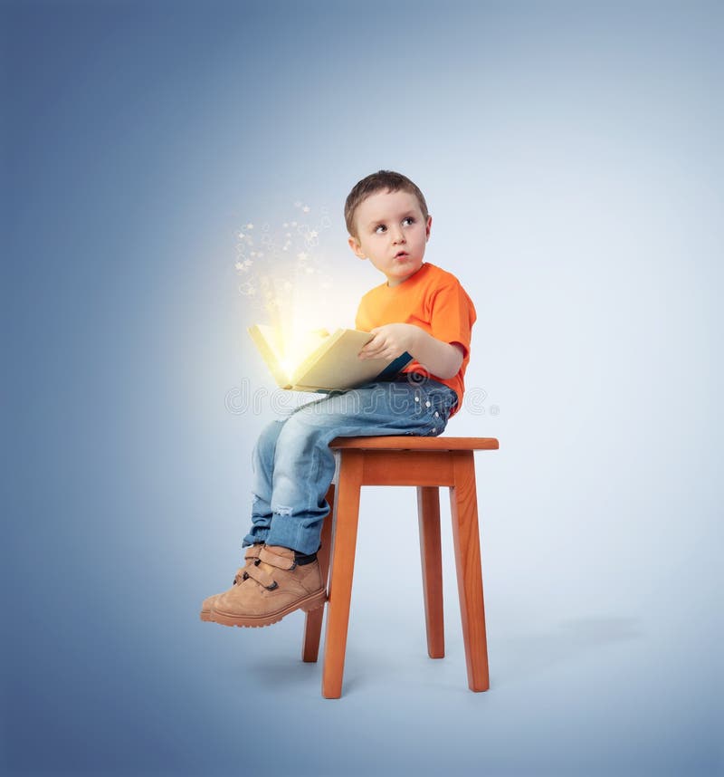 Ragazzino che si siede su una sedia con un libro magico aperto, su fondo blu Concetto di fiaba immagini stock