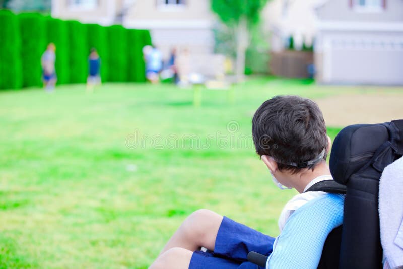 Ragazzino disabile nel gioco di bambini di sorveglianza della sedia a rotelle su gioco immagine stock Ragazzino disabile nel gioco di bambini di sorveglianza della sedia a rotelle su gioco immagine stock