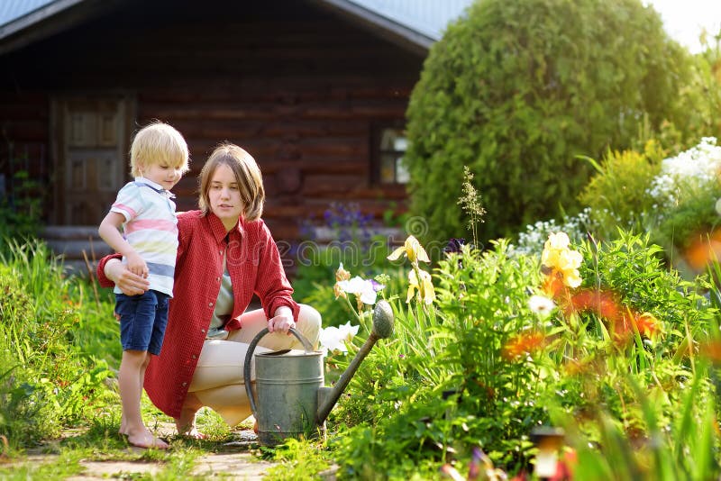 Ragazzino sveglio e le sue giovani piante di innaffiatura della madre nel giardino al giorno soleggiato di estate fotografia stock libera da diritti Ragazzino sveglio e le sue giovani piante di innaffiatura della madre nel giardino al giorno soleggiato di estate fotografia stock libera da diritti