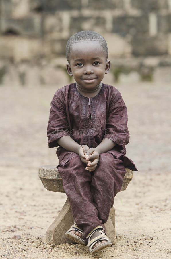 Ragazzo africano che si siede sul banco di legno e che esamina macchina fotografica con fondo vago immagine stock Ragazzo africano che si siede sul banco di legno e che esamina macchina fotografica con fondo vago immagine stock