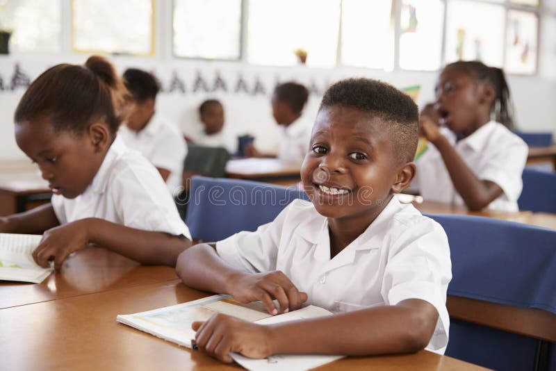 Ragazzo della scuola elementare che sorride alla macchina fotografica al suo scrittorio nella classe fotografia stock Ragazzo della scuola elementare che sorride alla macchina fotografica al suo scrittorio nella classe fotografia stock