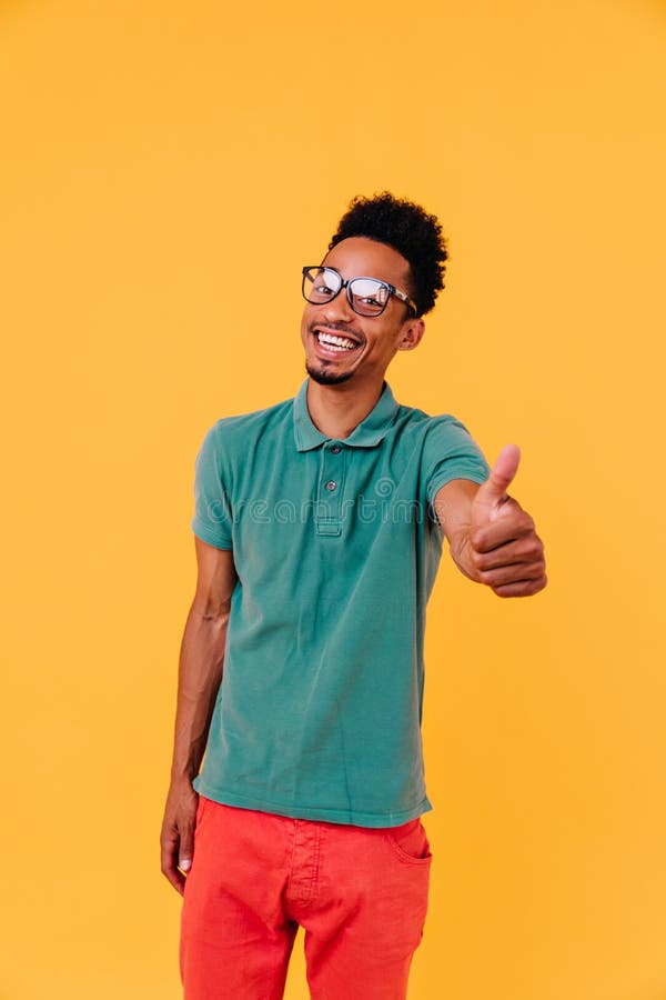 Ragazzo di buon umore in bicchieri grandi in posa con pollice su. foto da studio di un modello africano di sesso maschile fotografie stock libere da diritti
