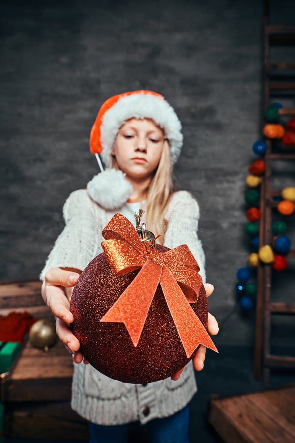 Ragazzo teenager che porta il cappello di Santa che tiene una grande palla di Natale in una stanza scura con una scala di legno d immagine stock libera da diritti Ragazzo teenager che porta il cappello di Santa che tiene una grande palla di Natale in una stanza scura con una scala di legno d immagine stock libera da diritti