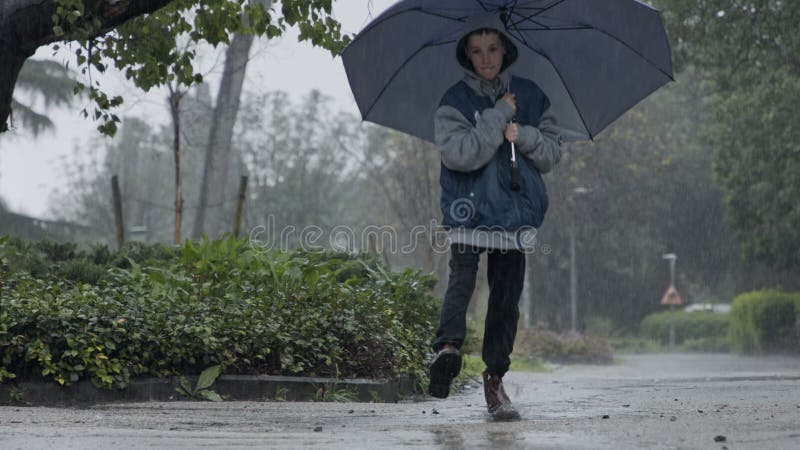 Ralenti d'un jeune garçon sautant sous une pluie battante tenant un parapluie clips vidéos