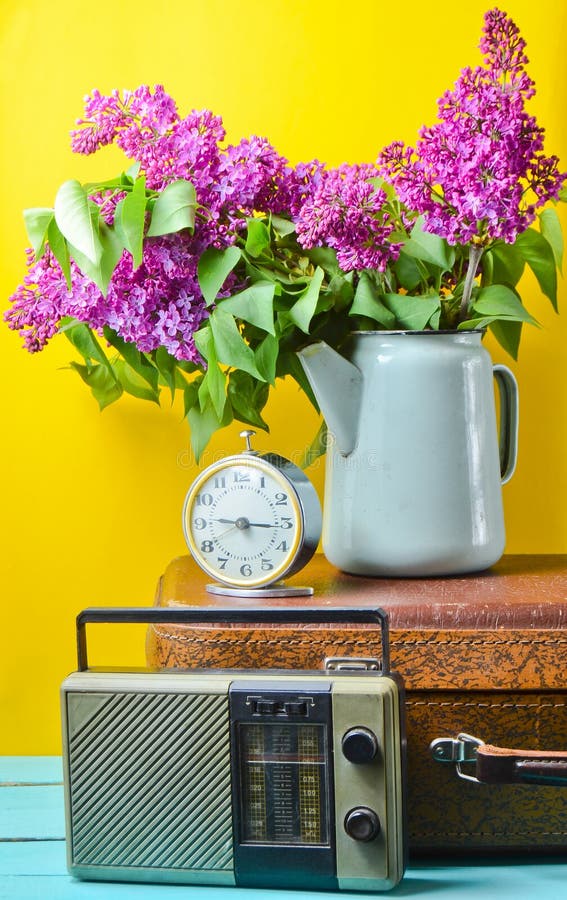 Ramalhete dos lilás na chaleira esmaltada na mala de viagem antiga, rádio do vintage, despertador no fundo amarelo Do estilo vida imagem de stock royalty free Ramalhete dos lilás na chaleira esmaltada na mala de viagem antiga, rádio do vintage, despertador no fundo amarelo Do estilo vida imagem de stock royalty free