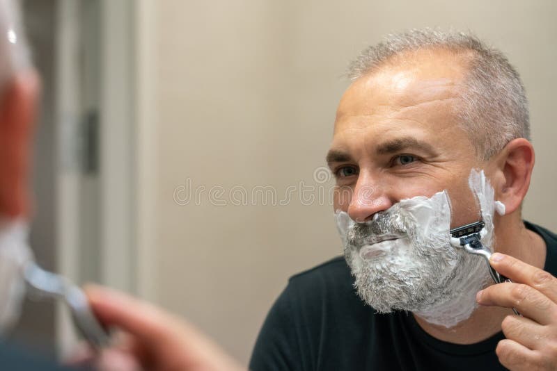 Handsome white-haired beared man shaving off his beard looking in mirror. Mature gray-haired man restyling his beard himself at home using razor. Handsome white-haired beared man shaving off his beard looking in mirror. Mature gray-haired man restyling his beard himself at home using razor
