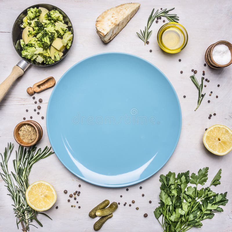 Raw broccoli in a small frying pan, parsley, oil, salt, lemon, pickles laid out around blue plate place for text,frame on wooden