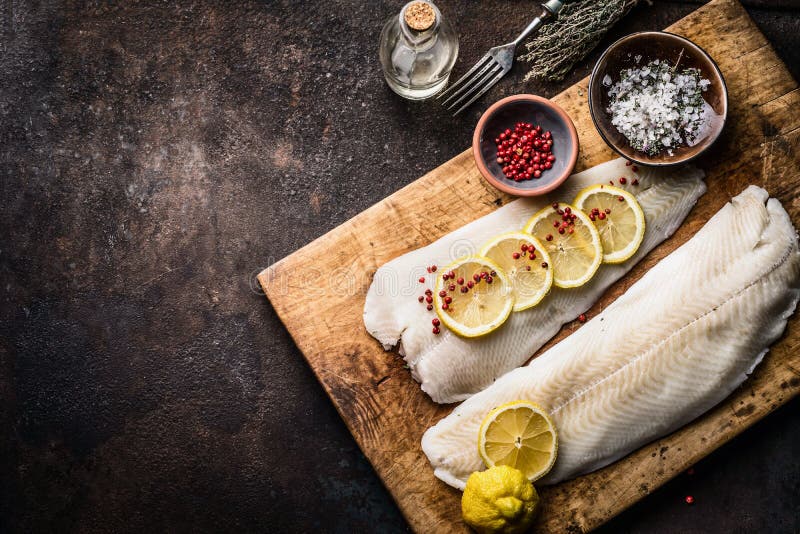 Raw cod fish fillet with lemon slices and herbal salt on rustic wooden cutting board on dark background, top view. Fish cooking