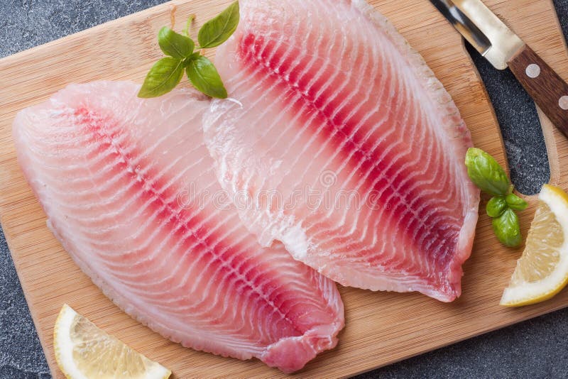 Raw fish fillet of tilapia on a cutting Board with lemon and spices. Dark table stock photos Raw fish fillet of tilapia on a cutting Board with lemon and spices. Dark table stock photos