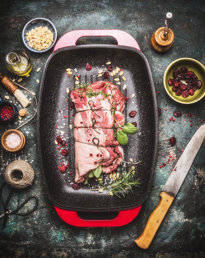 Raw Meat roast preparation in roaster pan with cooking ingredients and seasoning on dark rustic background with butcher knife