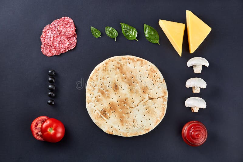 Raw pizza dough with ingredients for cooking vegetarian pizza on the dark stone surface with border, top view.