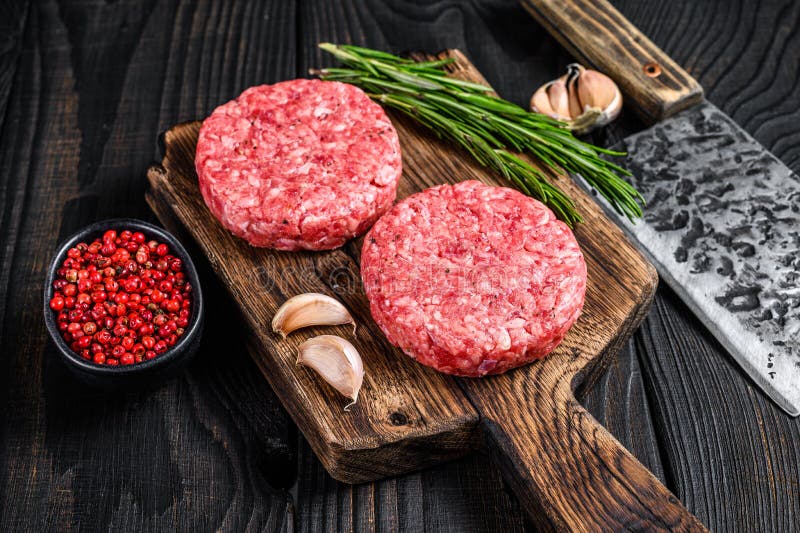 Raw steak cutlets with mince beef meat and rosemary on a wooden cutting board with meat cleaver. Black Wooden background