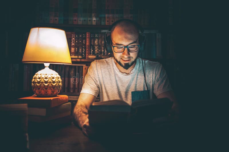 Reading and education. A man in headphones at the library reading a book in the evening by the light of a lamp. In the background stock photography Reading and education. A man in headphones at the library reading a book in the evening by the light of a lamp. In the background stock photography