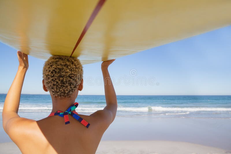 Rear view of African american woman in bikini carrying the surfboard on her head at beach in the sunshine