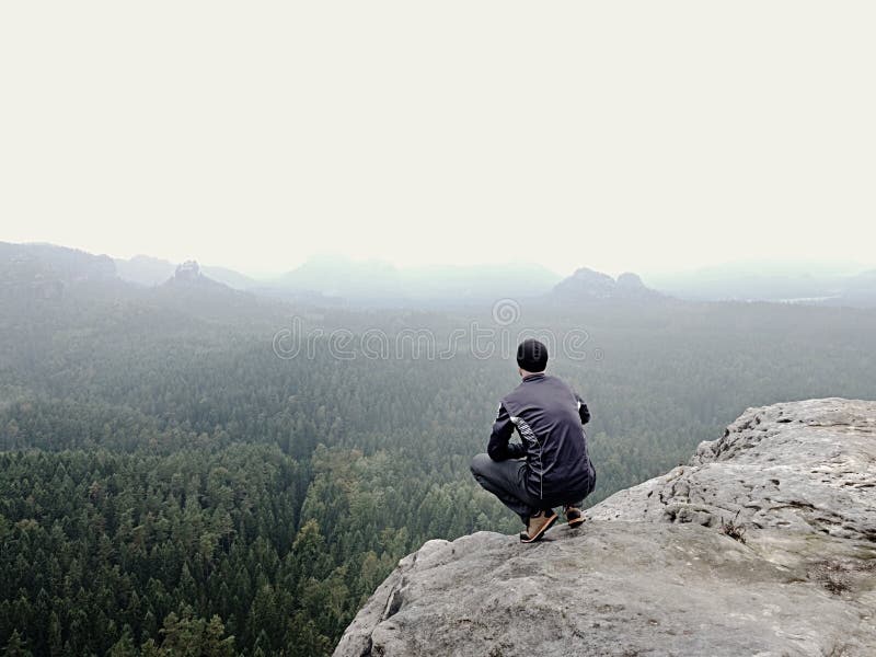 Rear view of alone hiker in dark outdoor suit sit on the edge. Sharp rocky peak above mountains valley, cold Sun hidden in rainy clouds.