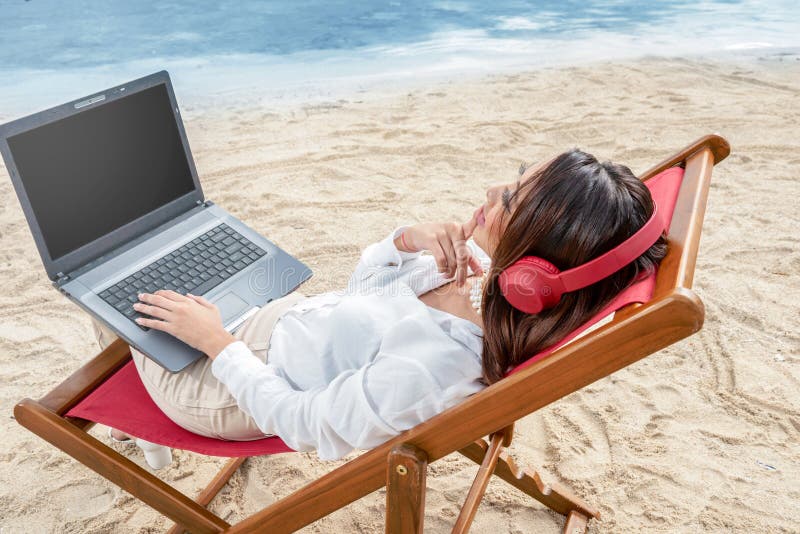 Rear view of asian business woman working with laptop while using headphones sitting in the beach chair and thinking something on