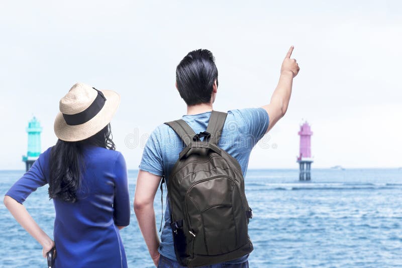 Rear view of Asian couple with hat and backpack looking at ocean view stock image
