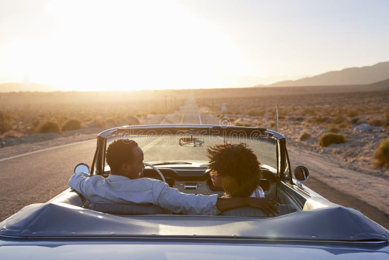 Rear View Of Couple On Road Trip Driving Classic Convertible Car Towards Sunset