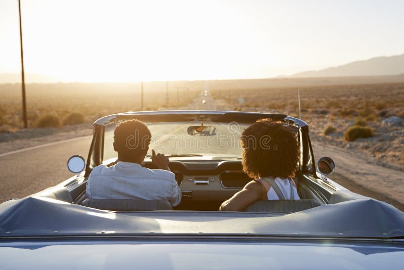 Rear View Of Couple On Road Trip Driving Classic Convertible Car Towards Sunset