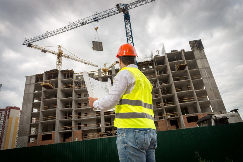 Rear view image of construction engineer in green safety vest and red hardhat controlling construction of new building