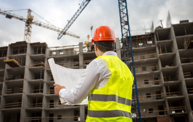 Rear view image of construction engineer looking at blueprints and working cranes on building site stock photo