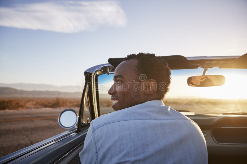 Rear View Of Man On Road Trip Driving Classic Convertible Car Towards Sunset