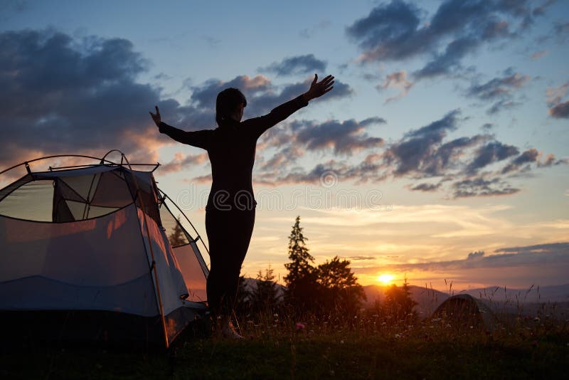 Rear view silhouette of female standing with open arms near camping in mountains at sunrise