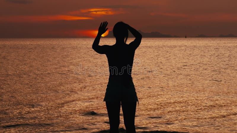Rear view silhouette of young woman looks at sunrise on sea and waves her hands greeting the sun rising from the horizon