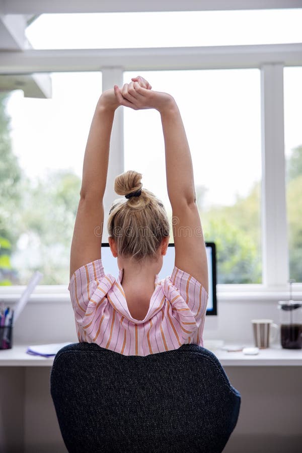 Rear View Of Woman Working From Home On Computer  In Home Office Stretching At Desk