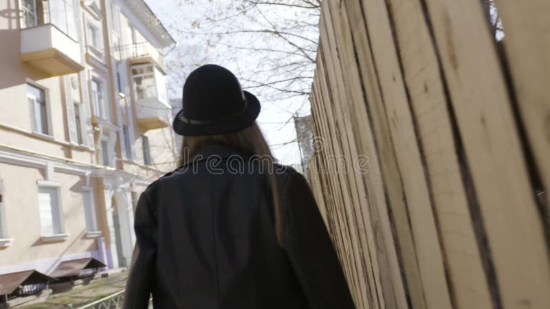 Rear view of a young tall girl in black leather jacket and a hat walking along the wooden fence in the street. Action