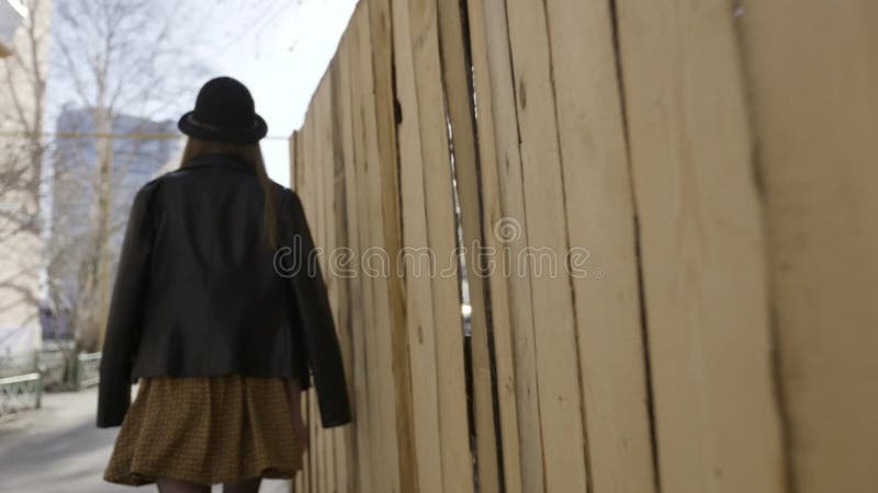 Rear view of a young tall girl in black leather jacket and a hat walking along the wooden fence in the street. Action