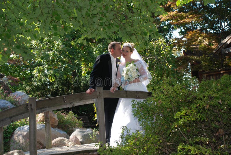 Groom kissing bride on cheek. Couple standing on a bridge in a park. Groom kissing bride on cheek. Couple standing on a bridge in a park.