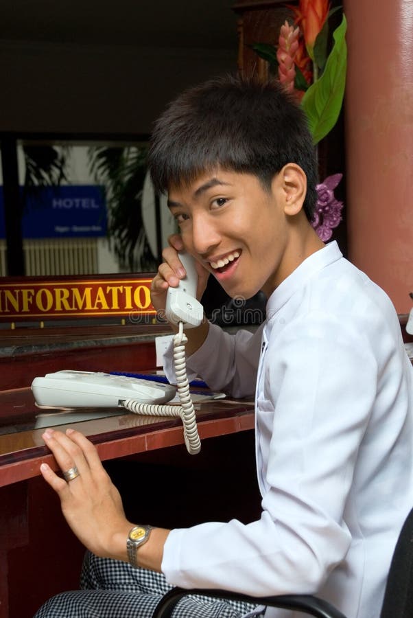 Asian smiling and welcoming hotel receptionist boy holding telephone hook or calling sitting at the desk of a hotel lobby with an information sign. Asian smiling and welcoming hotel receptionist boy holding telephone hook or calling sitting at the desk of a hotel lobby with an information sign.