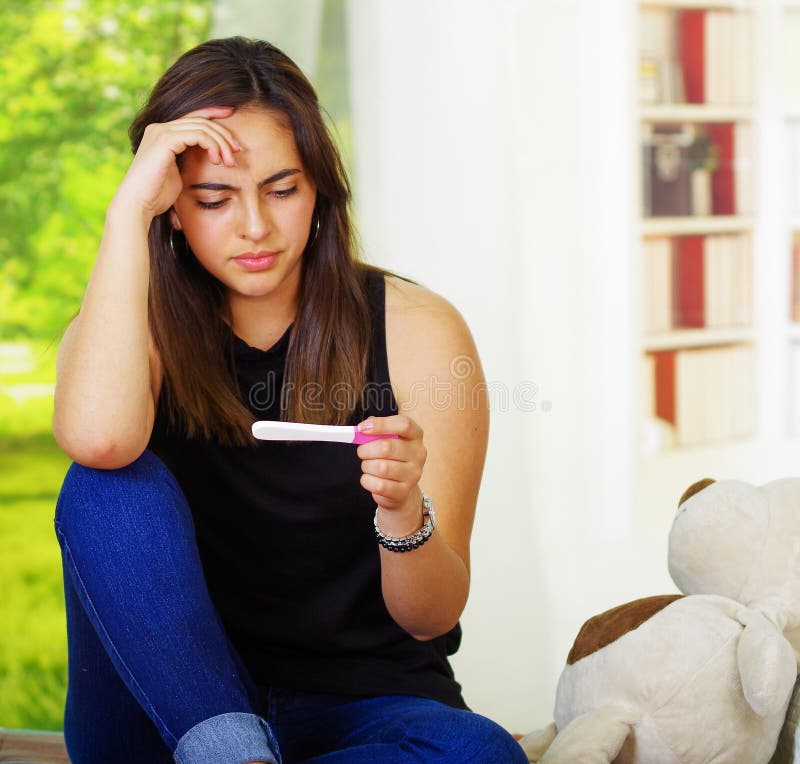 Recht junge Brunettefrau, die Schwangerschaftshaupttest, rührenden Kopf mit der Hand hält und im Unglauben, Bücherregale schaut stockfoto Recht junge Brunettefrau, die Schwangerschaftshaupttest, rührenden Kopf mit der Hand hält und im Unglauben, Bücherregale schaut stockfoto