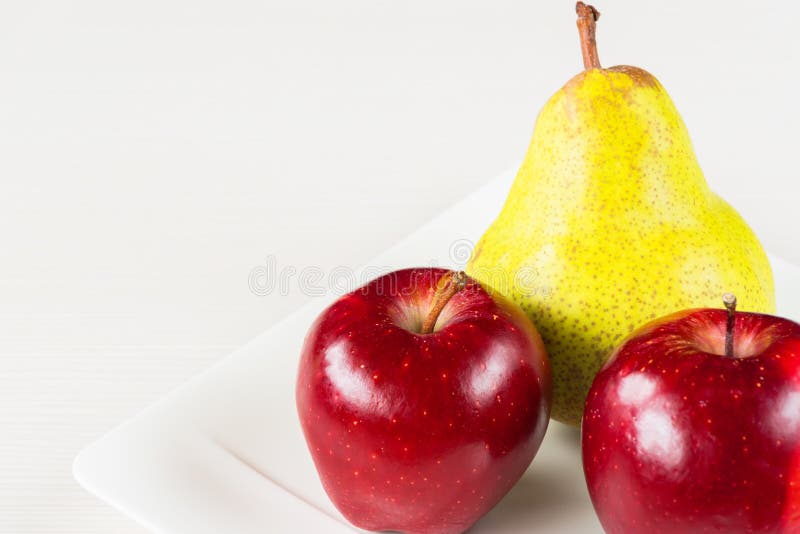 Red apples, green pear in white plate on white wooden background. stock image