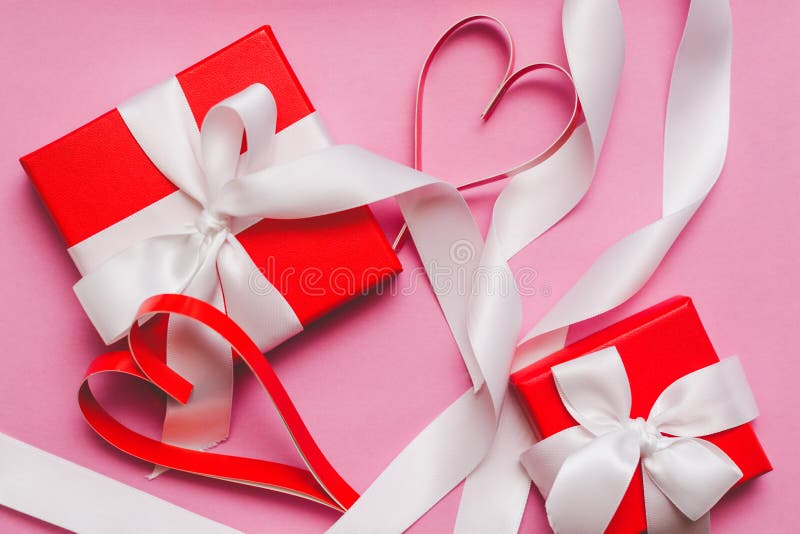 Red boxes with a gift, tied with a white ribbon, and red homemade paper hearts on a pink background. Symbol of Valentine`s Day.