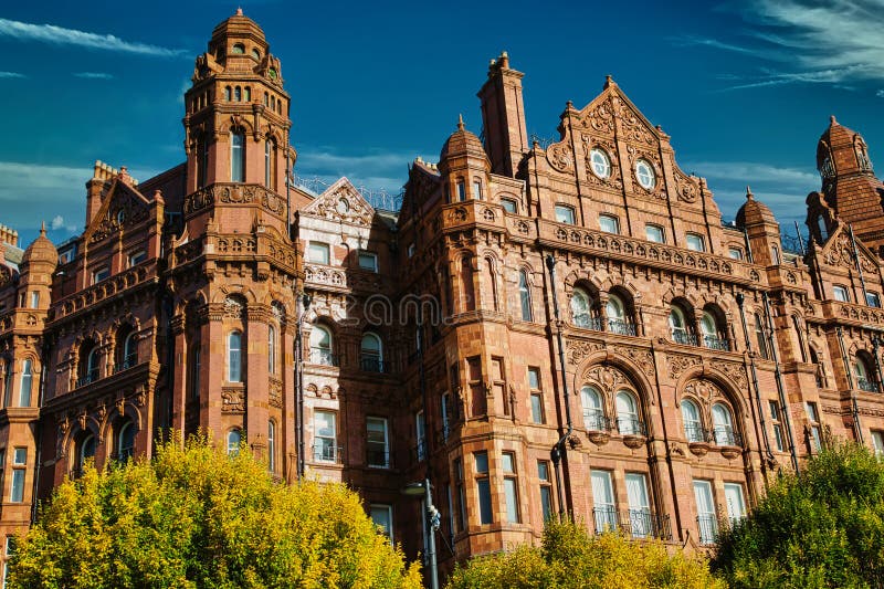 Red Brick Building with Ornate Details and Clock Face. A red brick building with intricate details, multiple windows, and a clock face, viewed from a low angle. Trees frame the building, partially obscuring the view. The sky is blue with some clouds
