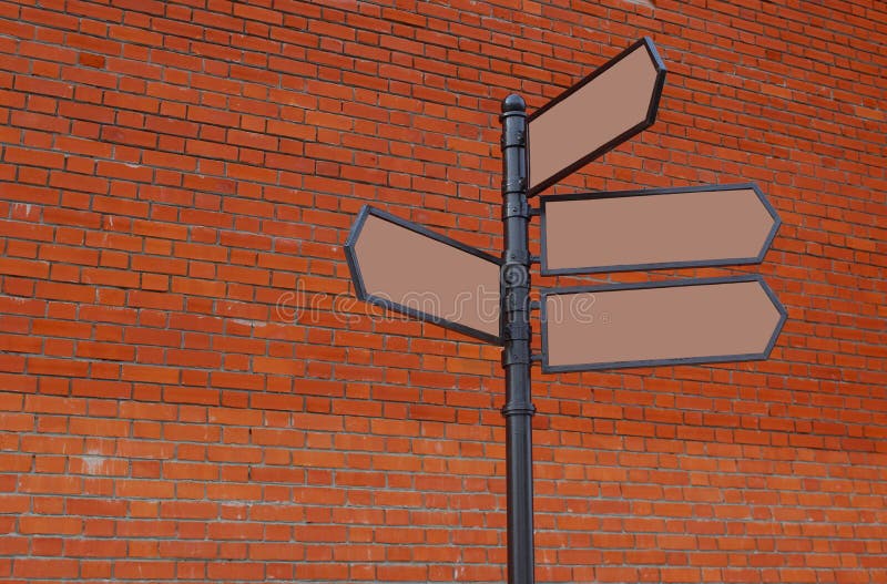 At the red brick wall is an iron column with arrows. Sign, post, pointer, signpost, direction, directional, road, arrow, blank, column, guide, travel, destination, information, signs, street, concept, arrows, tourism, metal, wood, banner, directions, guidepost, crossroad, symbol, crossroads, way, location, navigation, wall, brick, red, silicate
