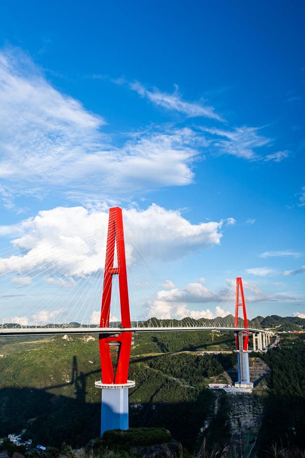 A striking red cable-stayed bridge spans between high mountains and canyons, with two prominent red pylons extending skyward. Its sleek structure is contrasted by the lush green forests below and a bright blue sky filled with scattered white clouds. The bridge links the elevated terrains, showcasing impressive engineering within a natural landscape. Red pylons stock images, royalty-free photos and pictures