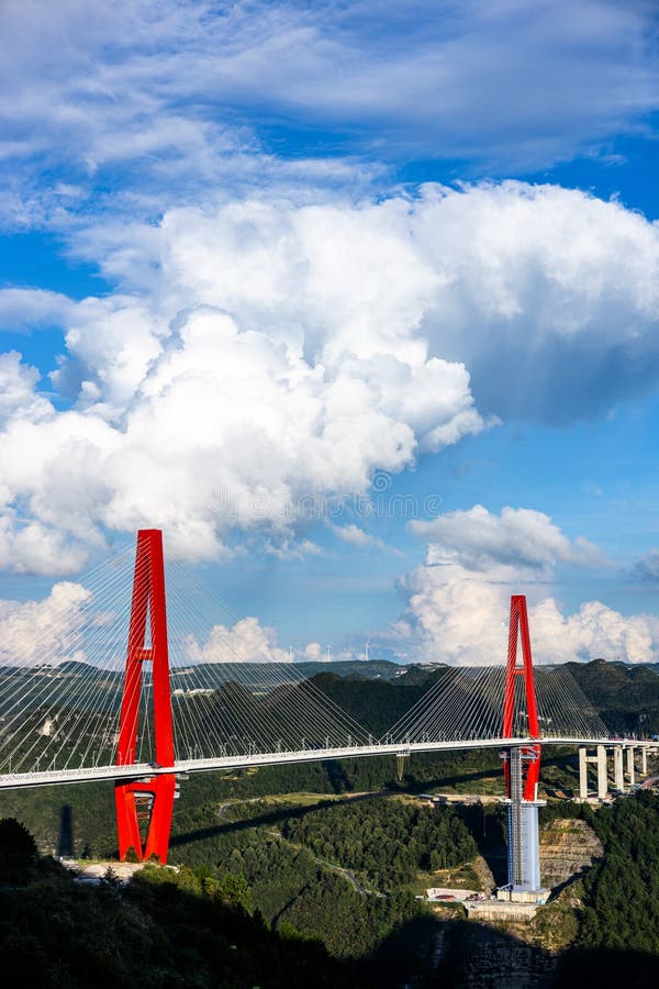 Tall red bridge with cable-stayed design spanning a deep valley in China. Surrounded by lush green hills, the bridge features two prominent pylons and connects over a forested area. Clear blue sky with large, fluffy white clouds provide a striking contrast to the bridge's vibrant color. The structure is an example of modern engineering against a scenic backdrop. Red pylons stock images, royalty-free photos and pictures
