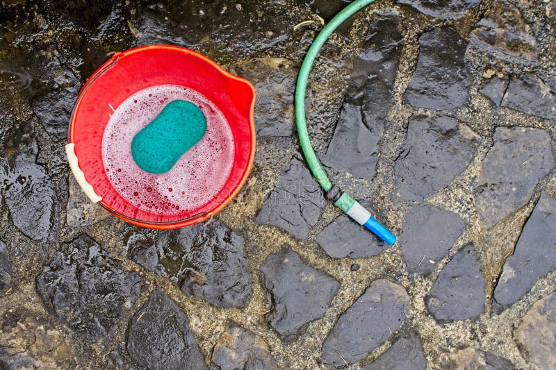 Red bucket with green sponge and soap on stone floor next to a green hose to clean cars