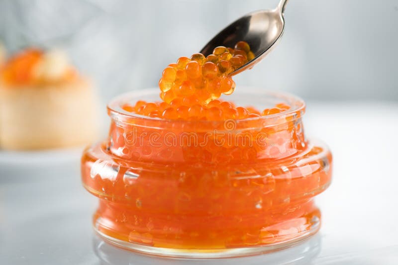 Red Caviar in a spoon, fish roe in a glass jar. Close-up of salmon fish roe caviar on served table. Delicatessen