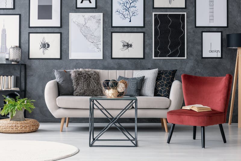 Red chair next to table and couch in modern apartment interior with gallery and plant on pouf. Real photo concept