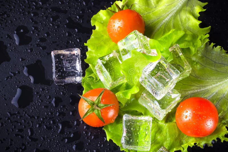 Red cherry tomatos, green salad and ice cubes on black wet table