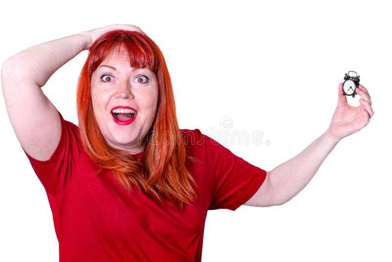 Red-haired middle aged woman wearing red T-shirt is holding a small alarm clock and screaming in shock. Concept of lack of time and being late. Isolated person. Red-haired middle aged woman wearing red T-shirt is holding a small alarm clock and screaming in shock. Concept of lack of time and being late. Isolated person