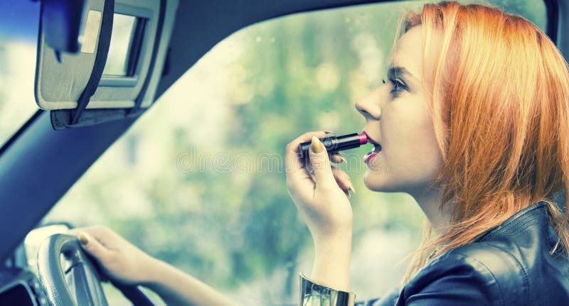Red haired young woman applying lipstick on lips in car. Beautiful girl making up inside. Danger on road and stereotypes. Red haired young woman applying lipstick on lips in car. Beautiful girl making up inside. Danger on road and stereotypes.